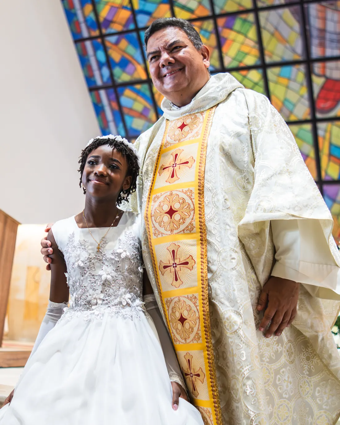 Un sacerdote del Santuario sonríe junto a una niña el día de su primera comunión, con el vitral detrás